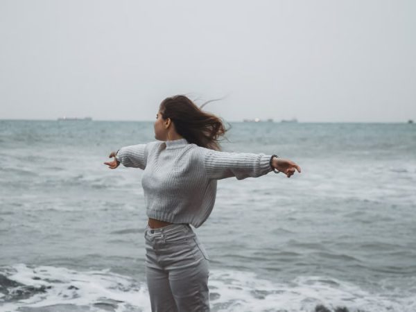 A woman stands with arms spread by the sea, embracing the freedom of a windy, overcast day.